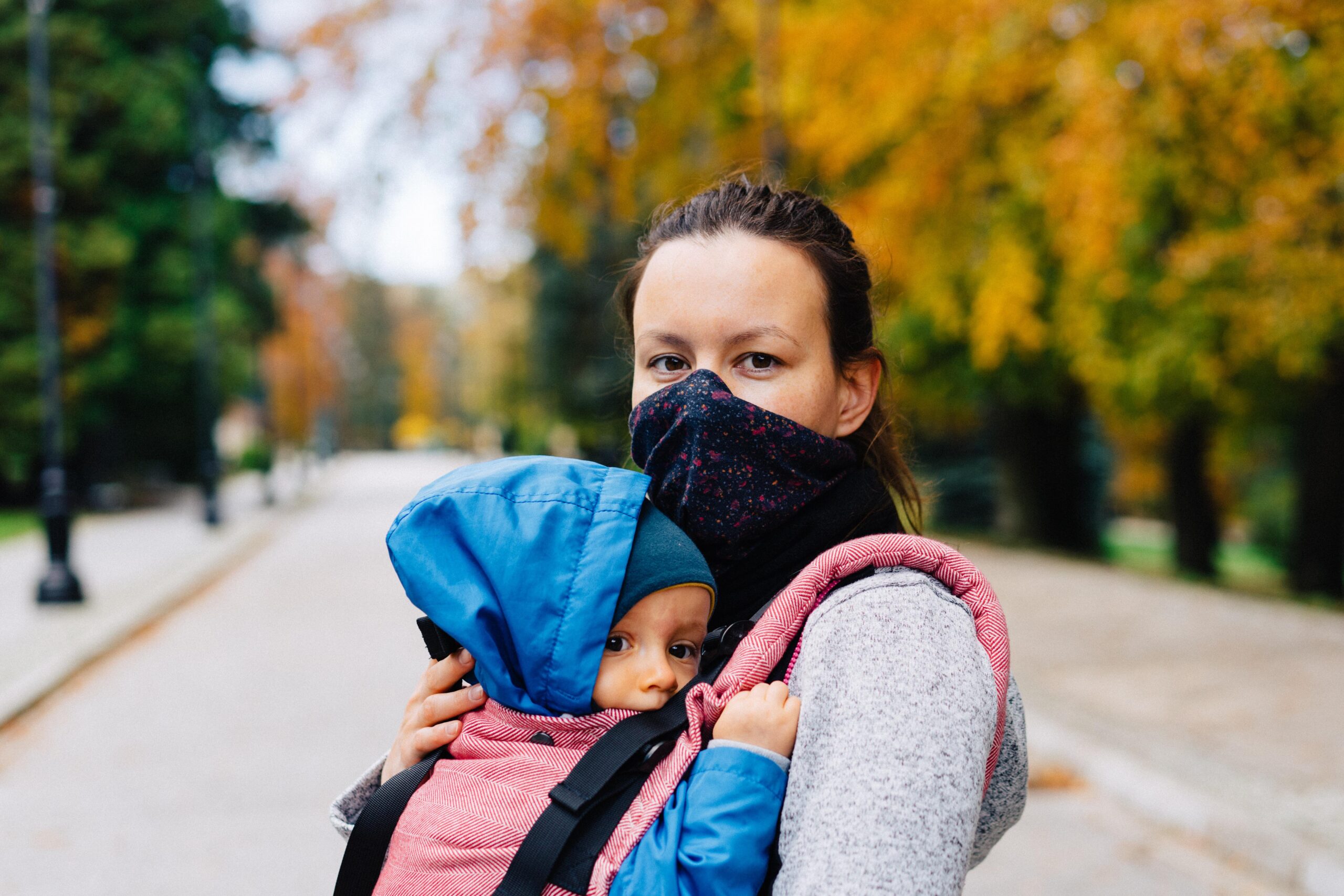 Woman with light skin and dark hair and facemask holding a baby in a baby carrier on their chest.